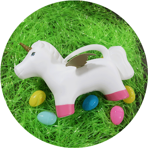 A white and pink unicorn shaped watering can, surrounded by green easter grass and colorful easter eggs.